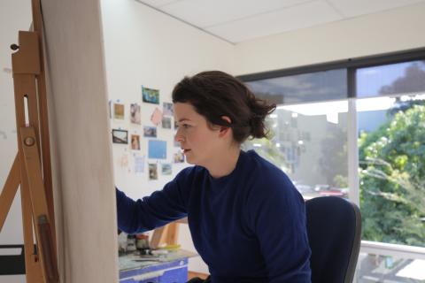 A photograph of a woman painting; she is wearing a blue sweater, and we are positioned behind her canvas.