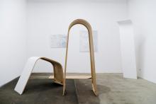 An installation view of abstract sculptures made of plywood, which appear to lean and bend on the floor and against walls, in a gallery space.