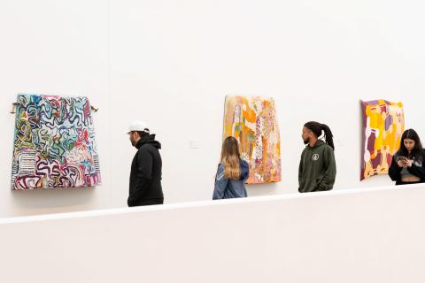 Three painted car bonnets are displayed on a white gallery wall, with visitors looking on
