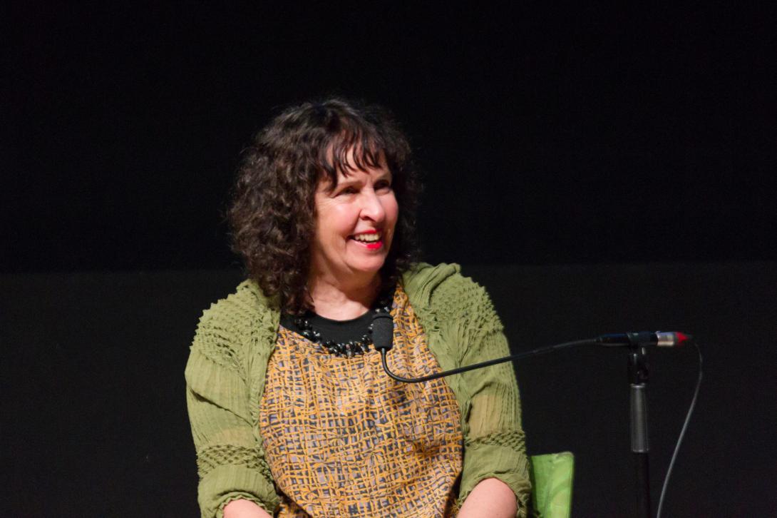 A photograph of a feminine-appearing person with wavy brown, shoulder-length hair, wearing a textured top and green cardigan, with a dark background; the person is smiling and about to speak into a microphone.