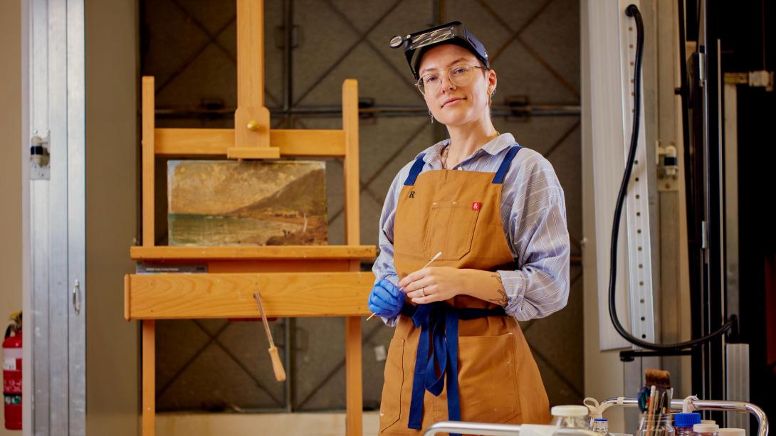 A conservator wearing an apron, glasses and with loupes propped up on their head stands proudly in front of a damaged oil painting they are about to begin restoring.