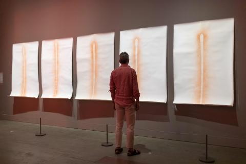 A visitor stands in a dark gallery space looking at a series of five works with an orange matchstick-like motif on a white background.