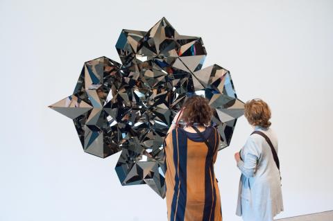 An installation view, with two women looking onto a shining, faceted metallic sculpture installed on a wall.