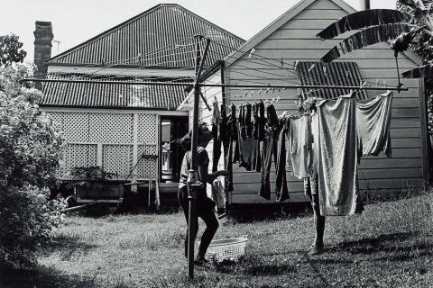 A black-and-white photo of a timber house, in front of which a person hangs washing on a Hill's Hoist-style washing line