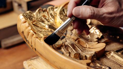 A conservation framer's hand holds a paintbrush over a frame mid-gilding