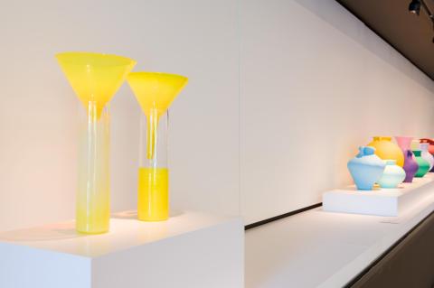 Sculptural works, including a neon yellow cylinder-and-funnel set and a colourful set of vases, installed in a gallery cabinet