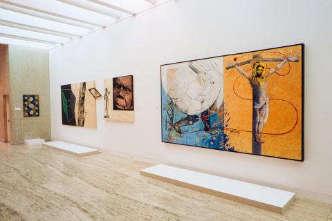An installation view of works installed on a white wall in a gallery space; there is one very large work at right that depicts a crucified Jesus, with other smaller works at left