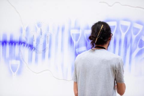 Facing away from us, an artist creates blue silhouettes of a spade handle on a white gallery wall in Reckitt's Blue paint