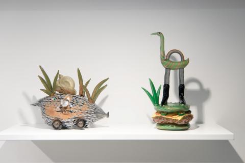 An installation view of two fanciful creatures made out of glass; one is half-car, half-carrot and the other is a tall bird standing on a burger.