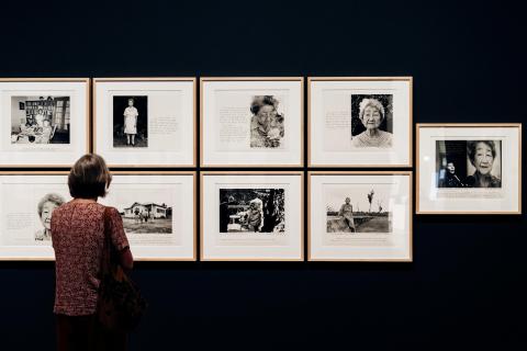 An installation view of black-and-white photographs installed on a dark gallery wall, with a person looking on