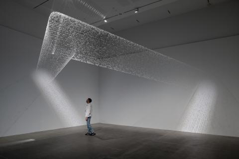 A view of a delicate spiderweb-like installation in a dimmed gallery space, with a visitor standing underneath the work.