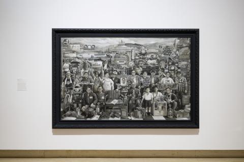 A large work rendered in black and grey tones installed on a white wall in a gallery space.