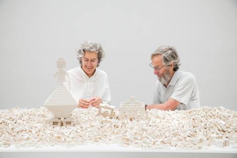 Two gallery visitors dressed in white construct architectural shapes from white Lego in a white gallery space 