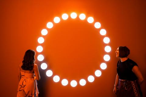 Drenched in red light, two gallery visitors gaze through small circular windows set in a circular formation into a gallery wall