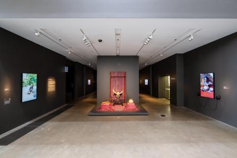An installation view of a small exhibition space with dark grey walls. An installation of red textiles is framed in the centre of the photograph, with video installations on walls at left and right.