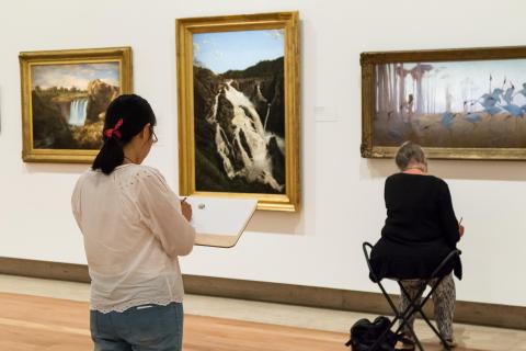 A photograph of two Gallery visitors sketching in the Australian Galleries; one stands further back from two paintings of waterfalls, which another sits on a fold-out chair near a painting of brolgas following a nude figure playing a flute.