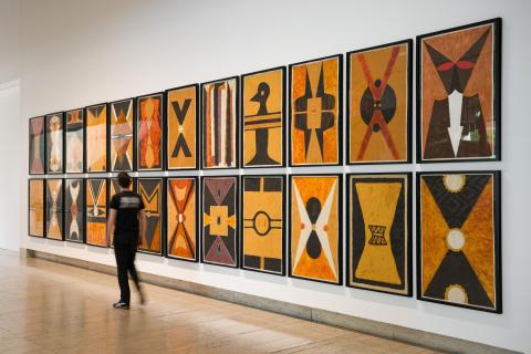A group of 24 'shield' paintings, using predominantly brown, tan and red pigments, installed in two rows on a white gallery wall. A man dressed in black walks in front of the installation. 