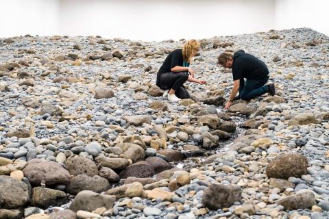 In this photograph, a person crouches on a rocky surface and extends a hand into a thin stream of running water that winds between grey and tan stones in a white-walled gallery.