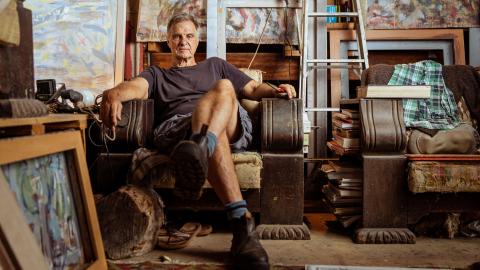 A photograph of a man sitting on a chair in his artist's studio, surrounded by works in progress, supplies and furniture.