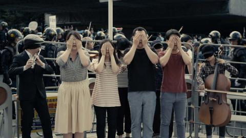 A photograph of four people covering their eyes; one person to the left plays flute and one to the right plays cello.