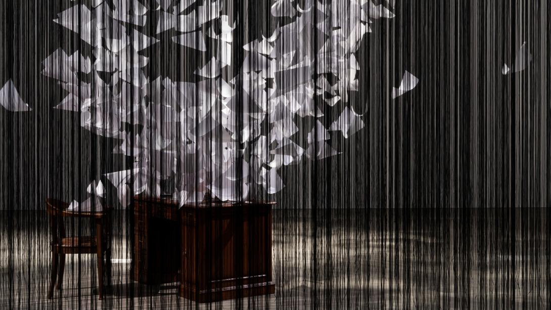 A detail view of a room-filling installation: seen through what appears as a curtain of black threads, a desk and empty chair have thousands of papers appearing to fly off of them in a tornado-like spiral. 