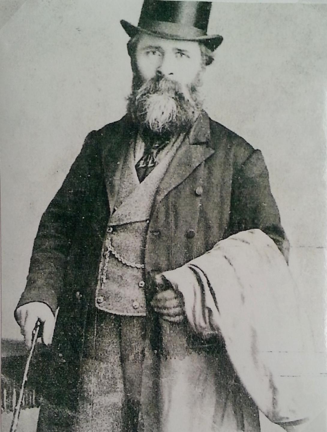 A black-and-white photograph of a man in the late 1800s; he is wearing a waistcoat and coat, a top hat, and carrying a cane, and looking directly into the camera.