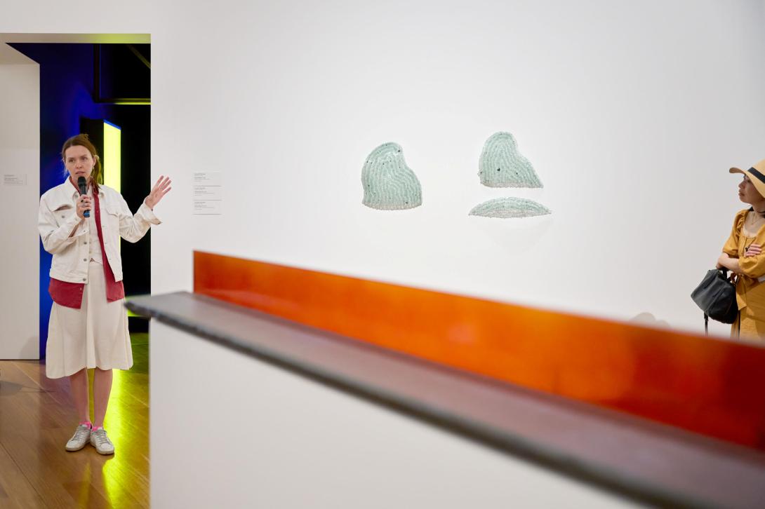 A photograph of an artist, at left, dressed in a red-and-white jacket and white skirt, speaking to a tour audience about the glass sculptural works installed on the rear wall; a large abstract sculpture is seen in the immediate foreground.