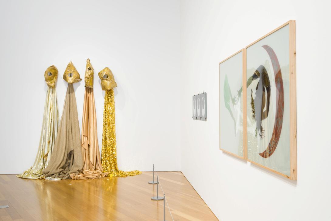 An installation view of a white gallery space with, at left, a gold-toned fabric work installed on the wall, with a diptych hung on the wall at right. 
