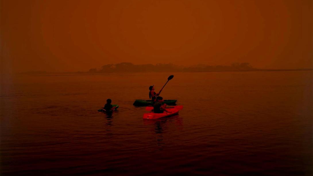 Rachel Mounsey / Australia b.1975 / Mallacoota fires in the sky 3 (detail) 2020 / Inkjet print on Canson Platine Fibre Rag paper / Purchased 2021. QAGOMA Foundation / © Rachel Mounsey A photograph of someone kayaking under a red post-bushfire sky.
