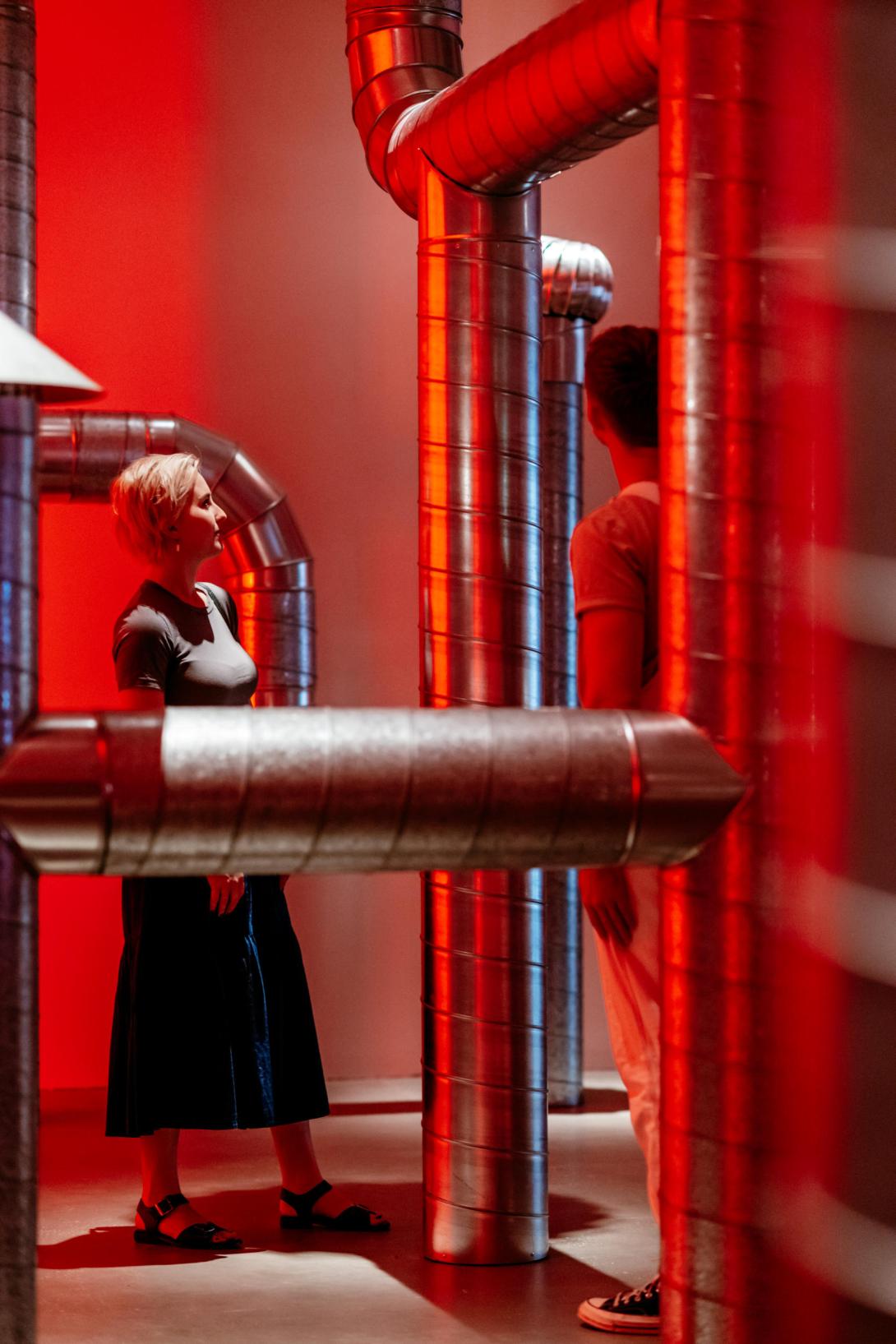 An installation view of a red-lit gallery space, with an artwork composed of piping criss-crossing the space.