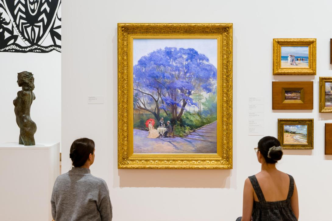 An installation view of a large oil painting depicting a jacaranda tree in bloom, surrounded by other artworks and with two visitors looking on from either side of the painting.