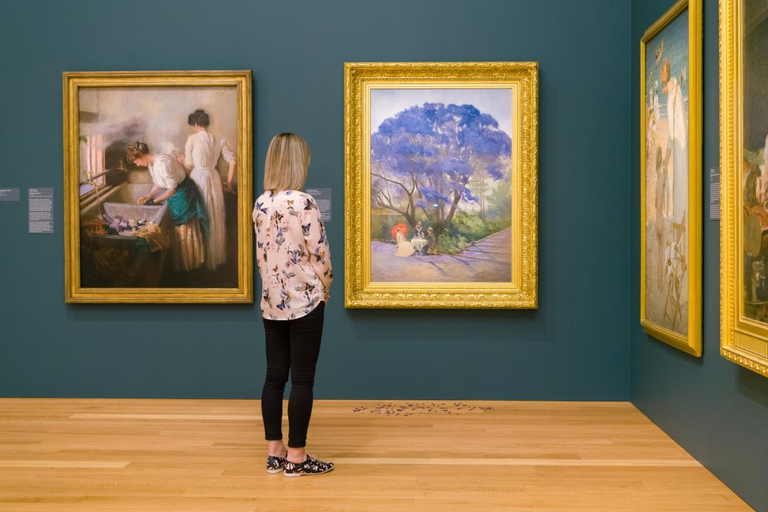 An installation view of a gallery with walls painted a dark blue-tinted green, with two large oil paintings; one depicts women washing laundry and the other depicts a large jacaranda tree. A visitor looks on.