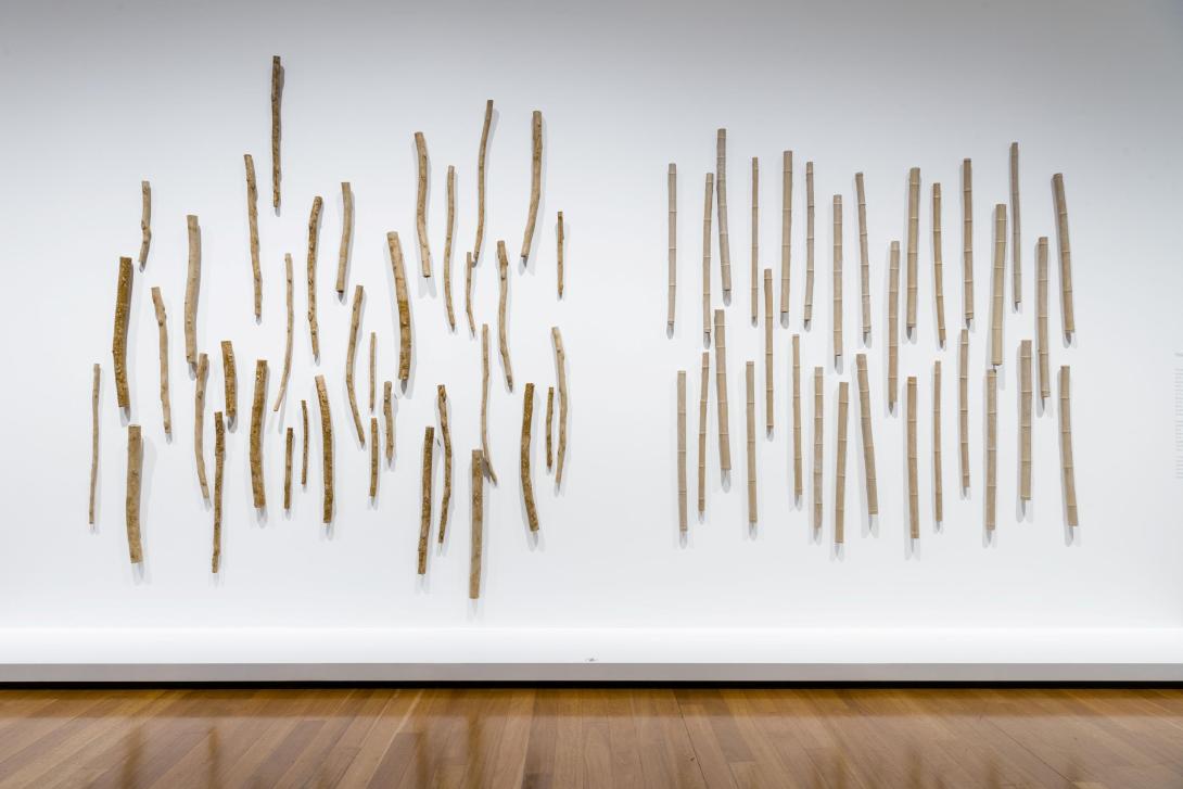 A sculptural work installed on a white gallery wall, taking the form of many gum and bamboo branches made from mud-coloured clay.