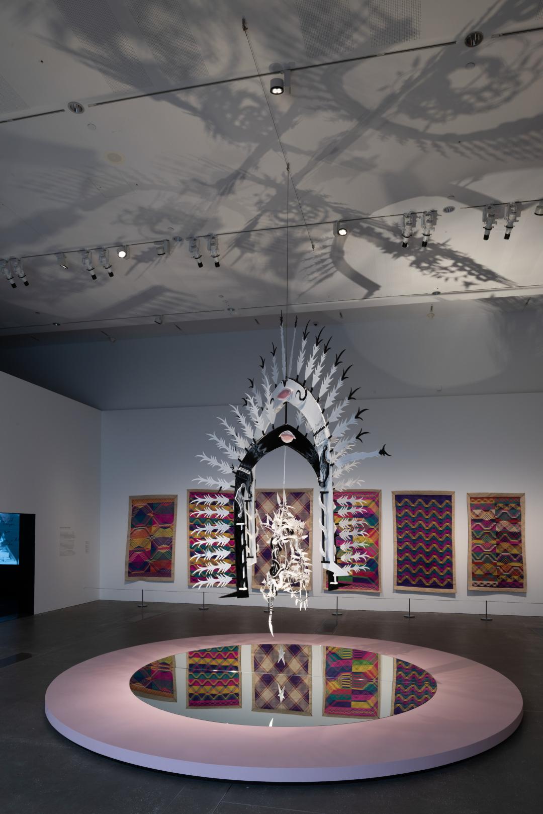 A tall kinetic dhari sculpture casts shadows onto the ceiling and reflected floor of a gallery space.