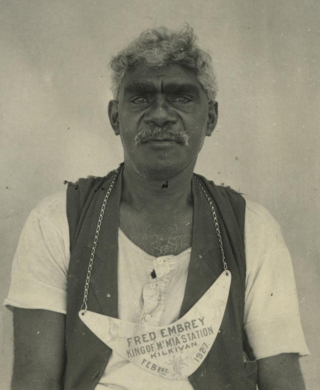 A black-and-white photograph of an Australian Indigenous man wearing a boomerang-shaped 'king plate' around his neck, along with a t-shirt and vest