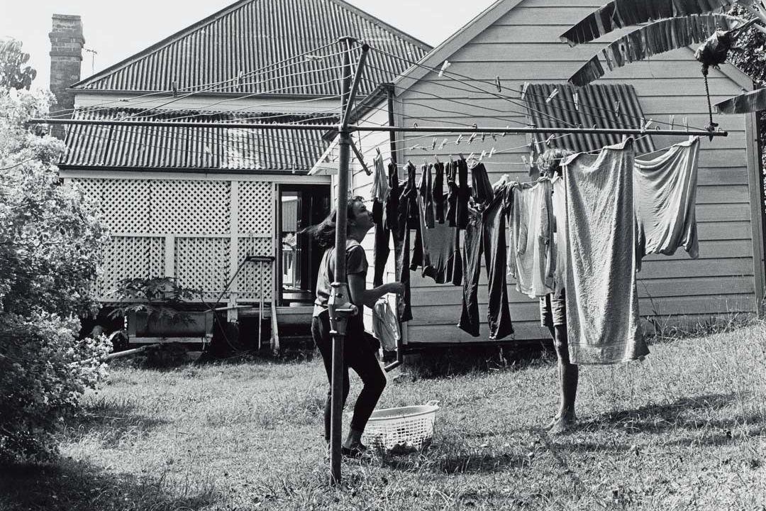 A black-and-white photograph of a person hanging clothes on a Hill's Hoist washing line in the backyard of a Queenslander-style house.