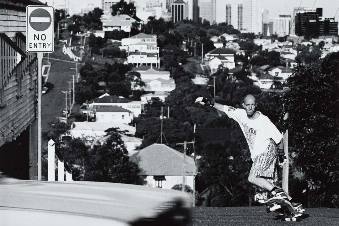 A black-and-white photograph of a man skateboarding on a steep suburban street, with houses and trees stretching out in the background, and a 'NO ENTRY' sign at left.