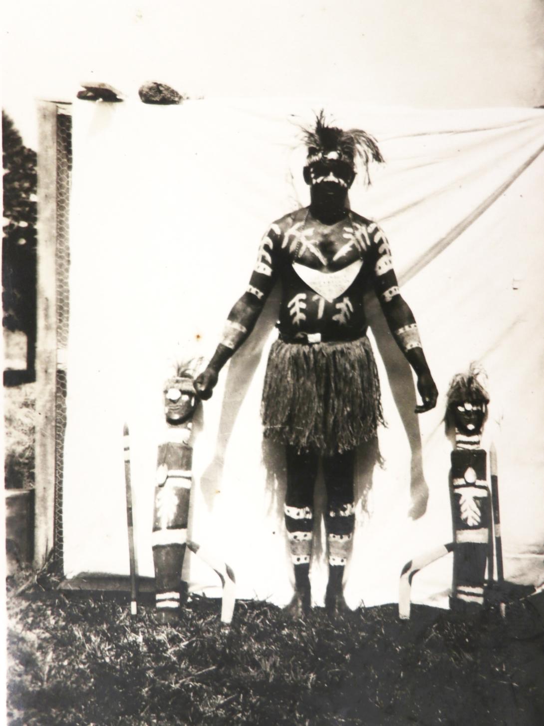 A black-and-white photograph of an Australian Indigenous man standing with two figurative sculptures