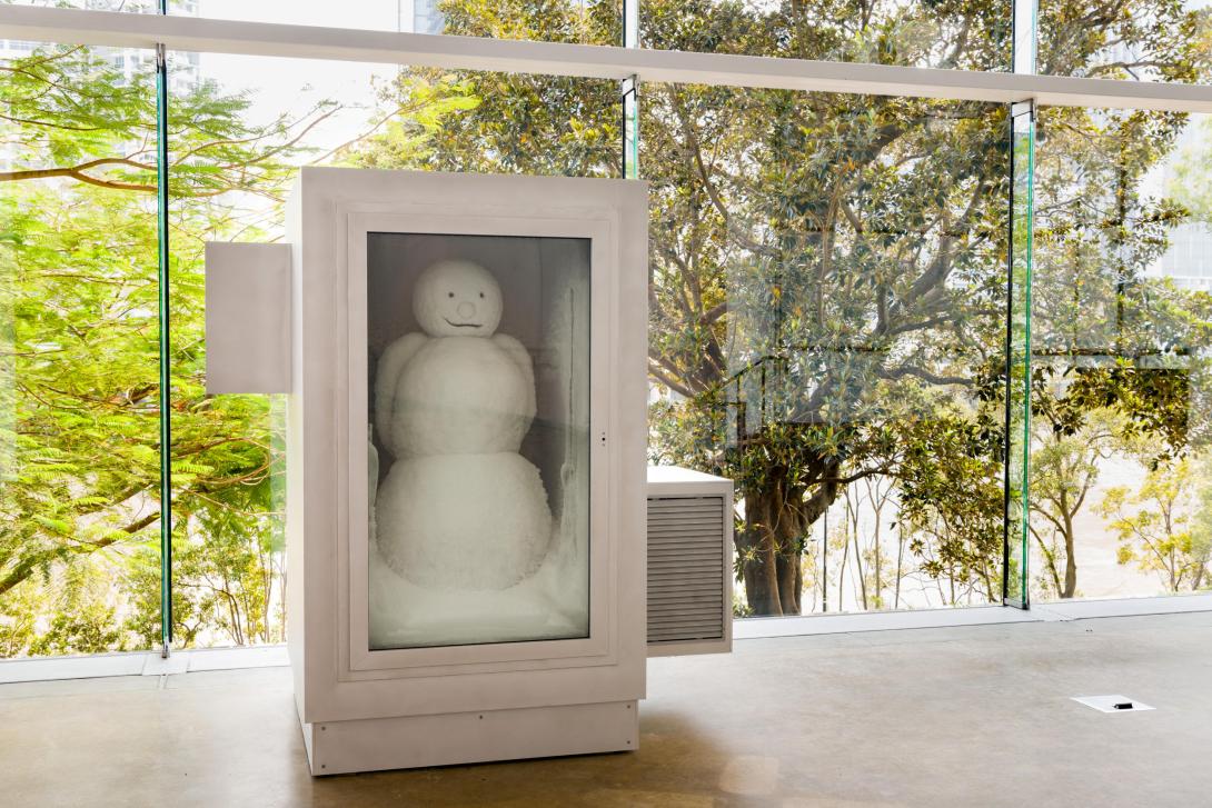 A freezer with a glass front is installed in a gallery space, in front of a glass wall looking over a green park; inside the freezer is a smiling snowman.