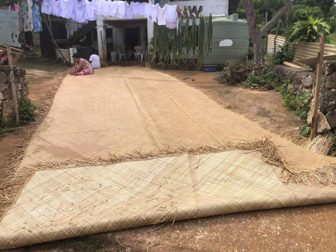 A photograph of a large mat outdoors, with two weavers working on it at the very end of the mat
