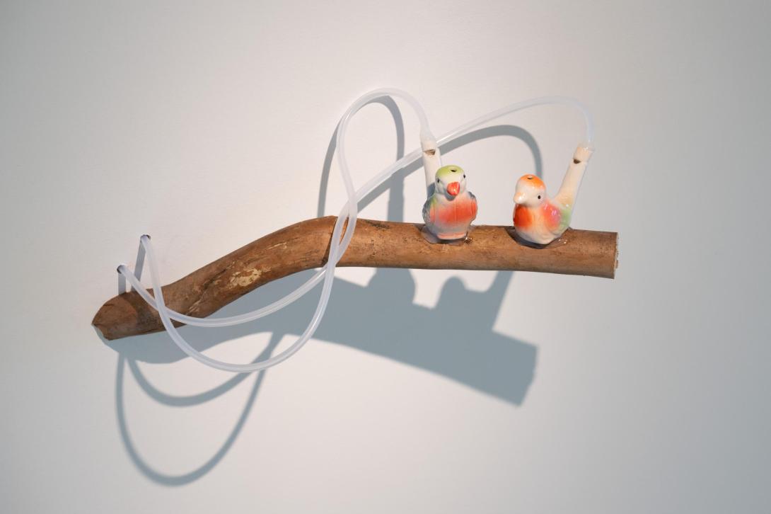 An artwork comprising a 'branch' and two small birds, with two white tubes attached, installed on a wall in a gallery space.