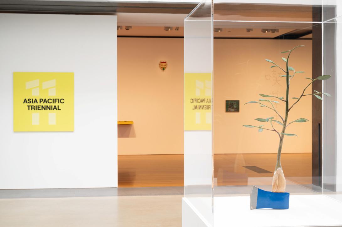 An installation view of a gallery space, with a sculpture that appears to be an axe with a sapling for a handle in the immediate foreground.