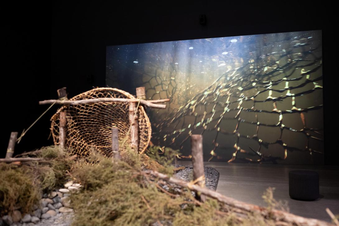 A fishing net and part of a river bed installed in a dark gallery space, with an underwater video behind
