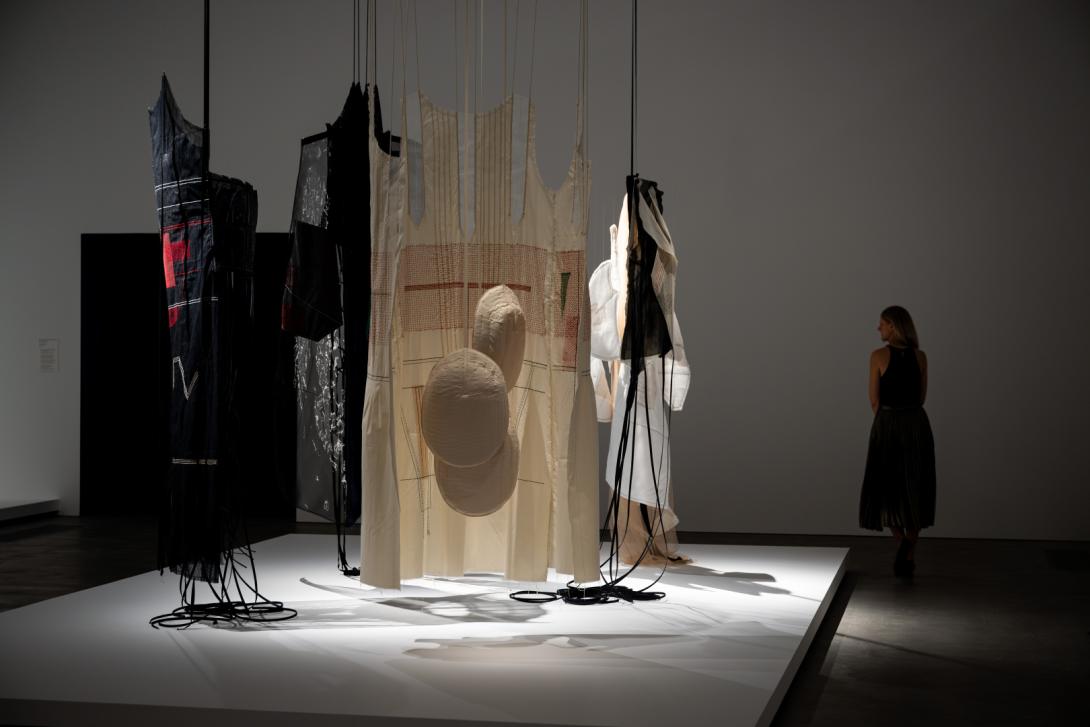 Suspended textile works evoking garments suspended over a plinth in a dimmed gallery space