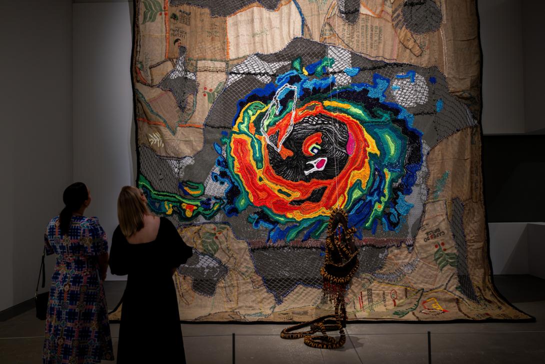 An installation view of a large, colourful textile work in a darkened gallery space, with two visitors looking on