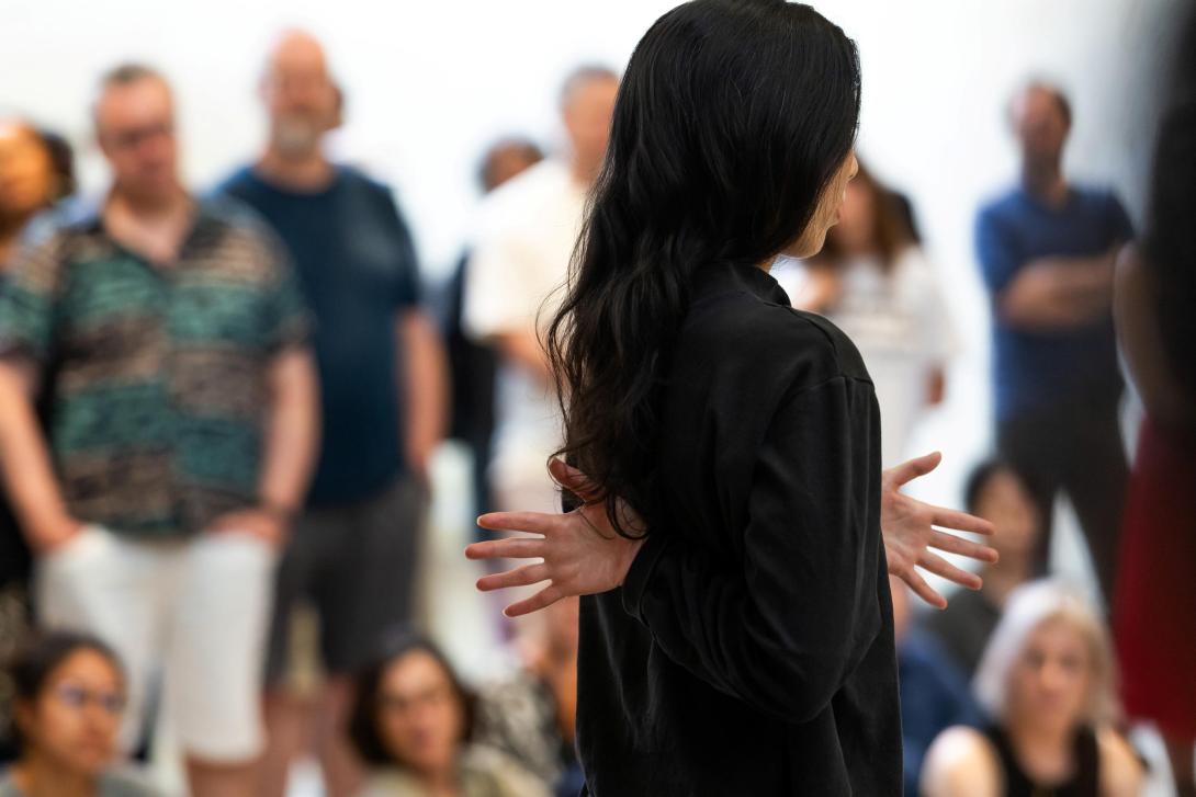 A dancer dressed in black, with long dark hair, performs midst a crowd of onlookers within a gallery space.