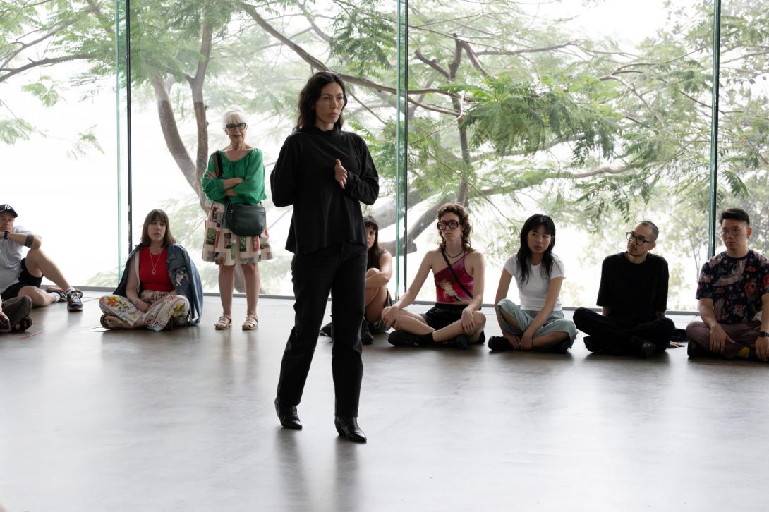 A dancer dressed in black, with long dark hair, performs midst a crowd of seated onlookers within a gallery space.