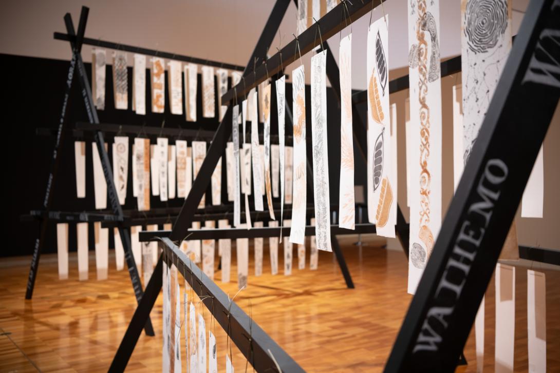Two wooden structures installed in a gallery space, with many long white sheets of paper suspended on the wooden frames