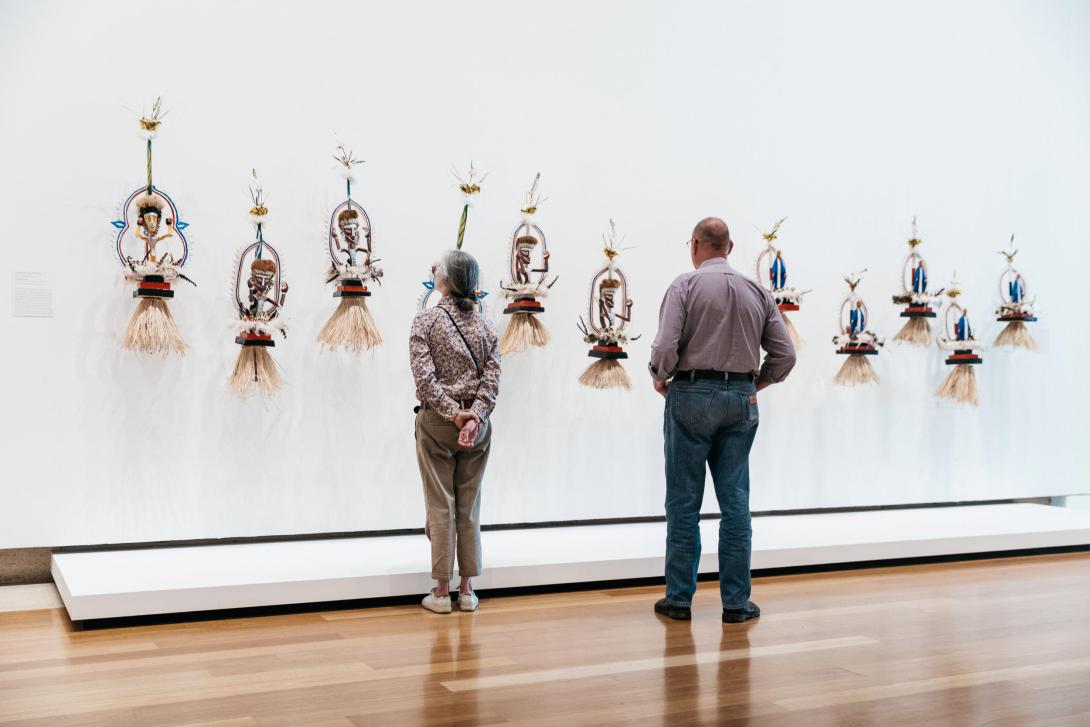 An installation view in which we see Papua New Guinean ceremonial headwear depicting various mythical characters, installed on a white gallery wall at eye-height; two visitors look on