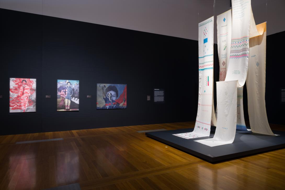 An installation view of a dimmed gallery space, with paintings installed on dark walls in the background and a large textile installation in the foreground